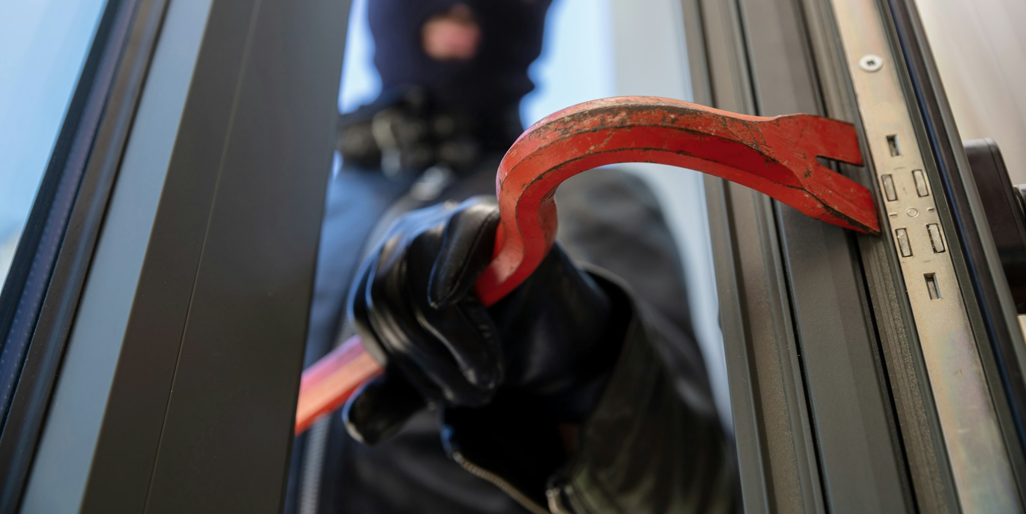 Bandit in balaclava holding a crowbar to break a glass window.