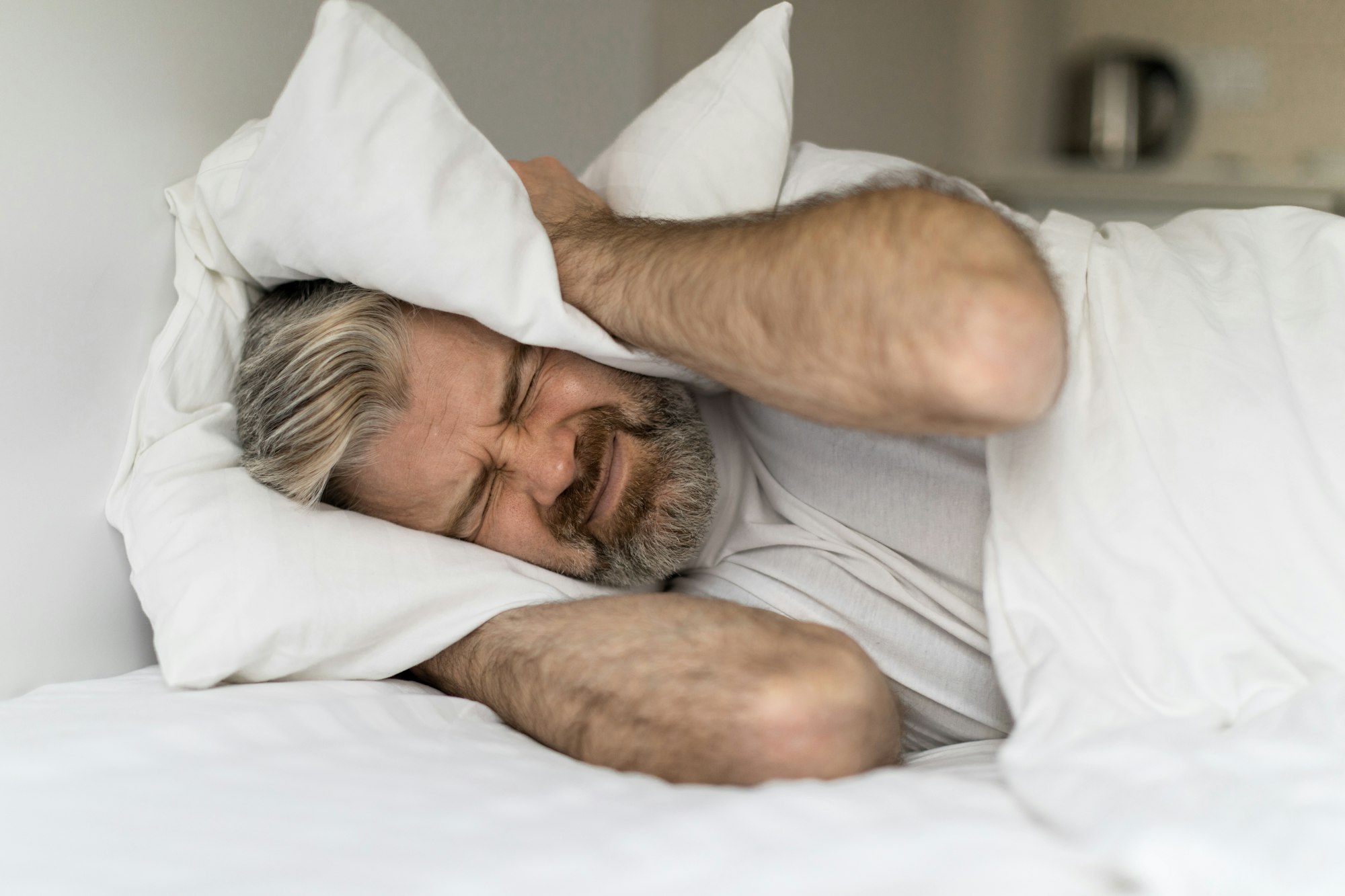 Irritated man lying in bed and covering ears with pillows